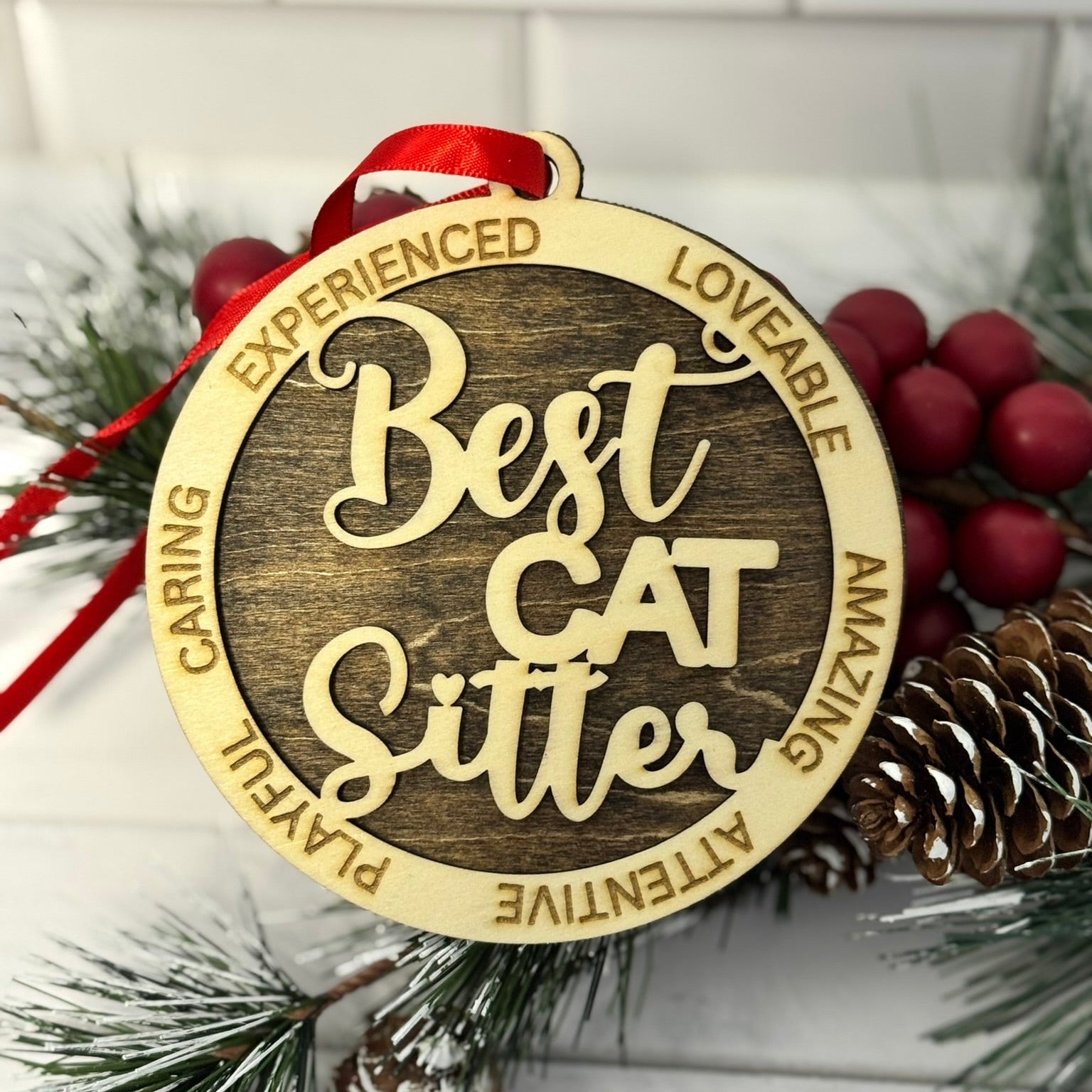 Decorative ornament with 'Best Cat Sitter' text on a wooden background with pine cones and berries.