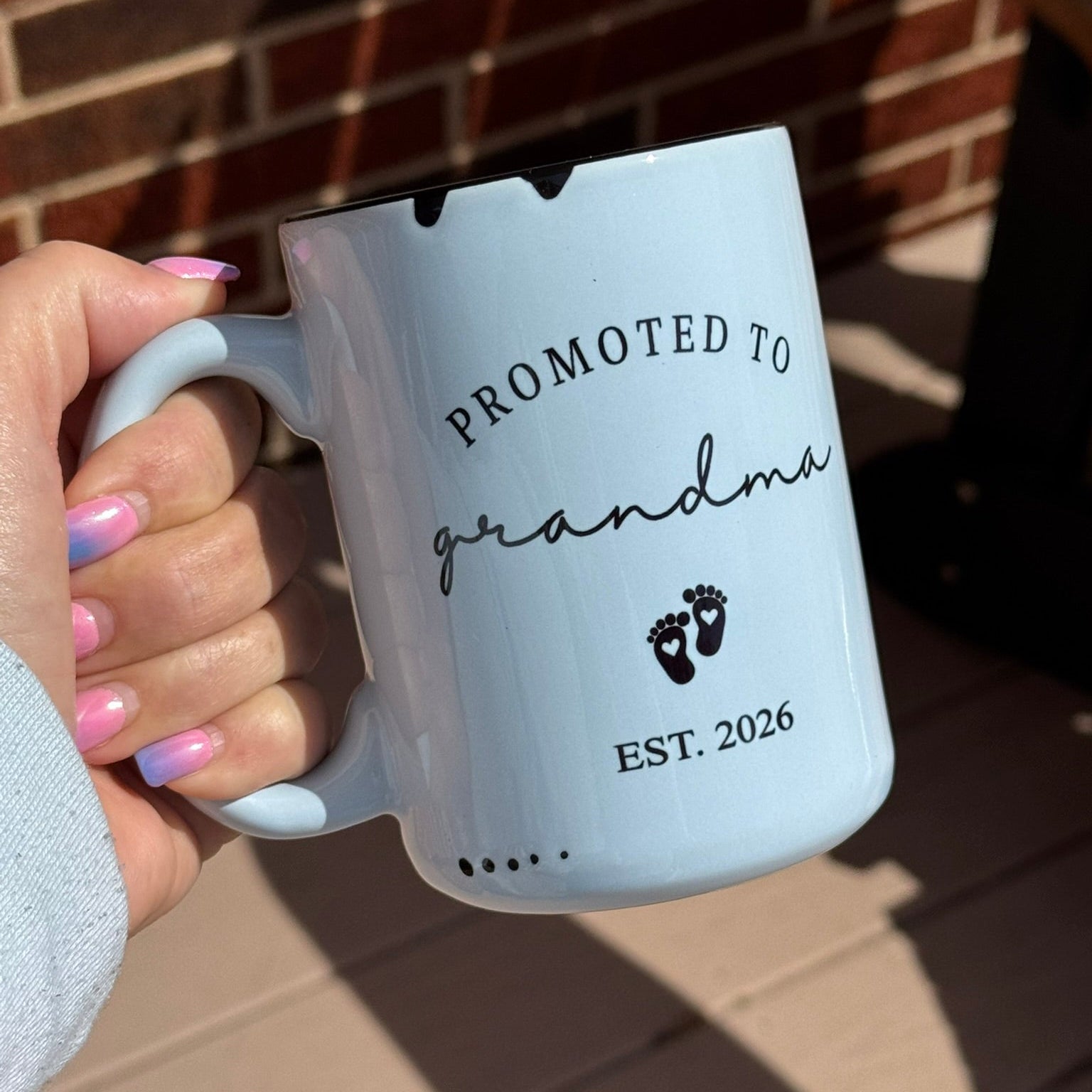 Light blue mug with 'Promoted to grandma' text held by a hand against a brick wall.